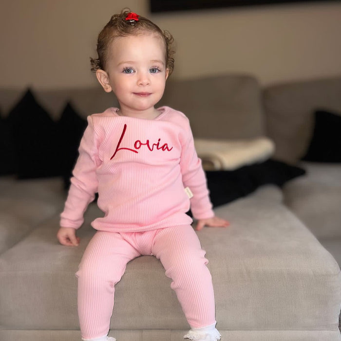 Little girl wearing a pink ribbed tracksuit with Lovia embroidered on the front in red