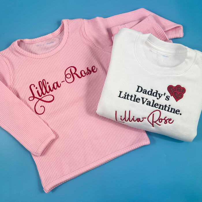 Pink ribbed tracksuit with name embroidered on the front in red and a white jumper folded next to it saying daddy’s little valentine