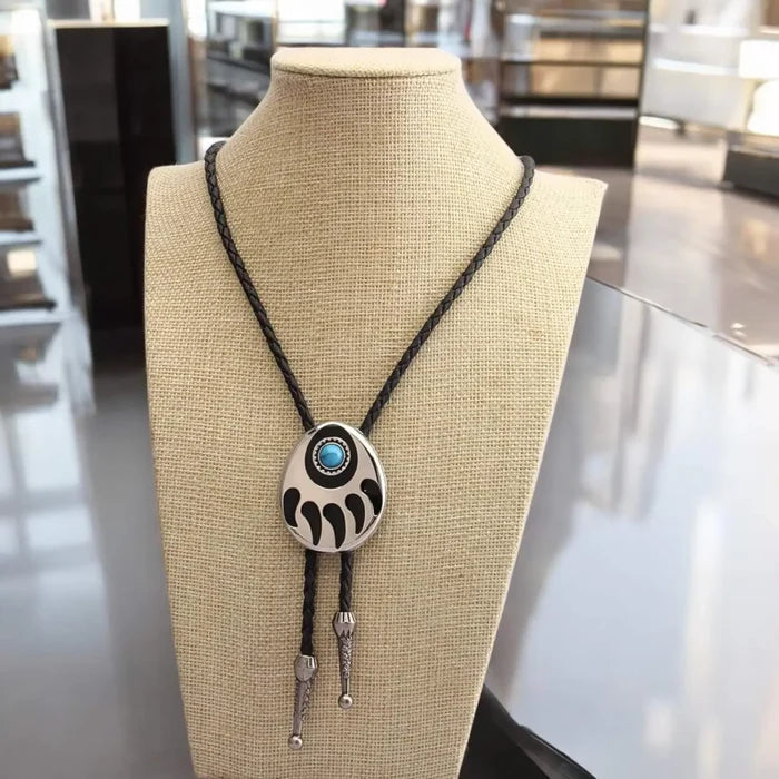 Two bear paw bolo ties with black braided leather cords: silver-tone faux turquoise bear claw slider and black stone bear paw pendant with antique metal tips.