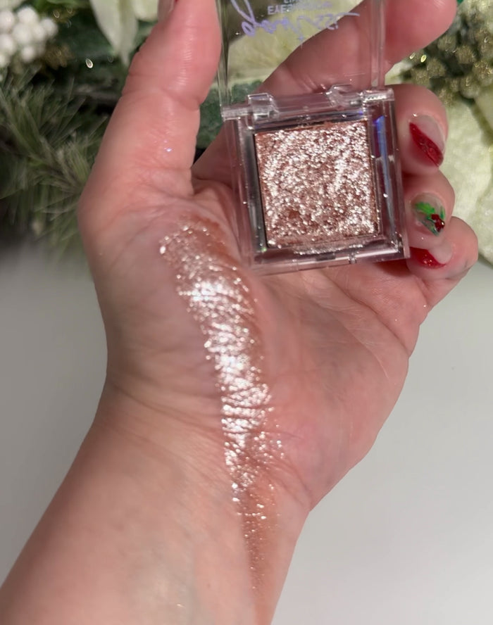 Hand holding shimmery eyeshadow compact and a swatch next to it, with a blurred floral background.