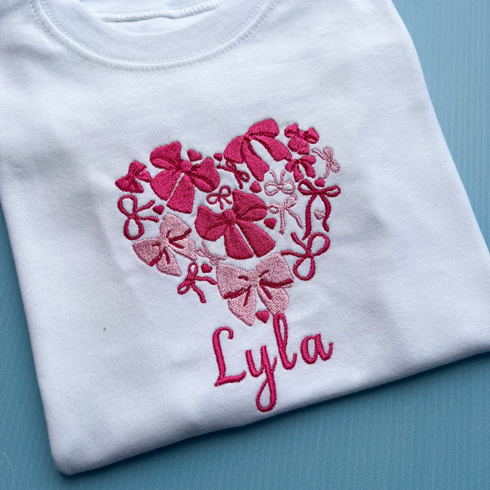 White shirt with pink embroidered heart design and name 'Lyla' on a blue background
