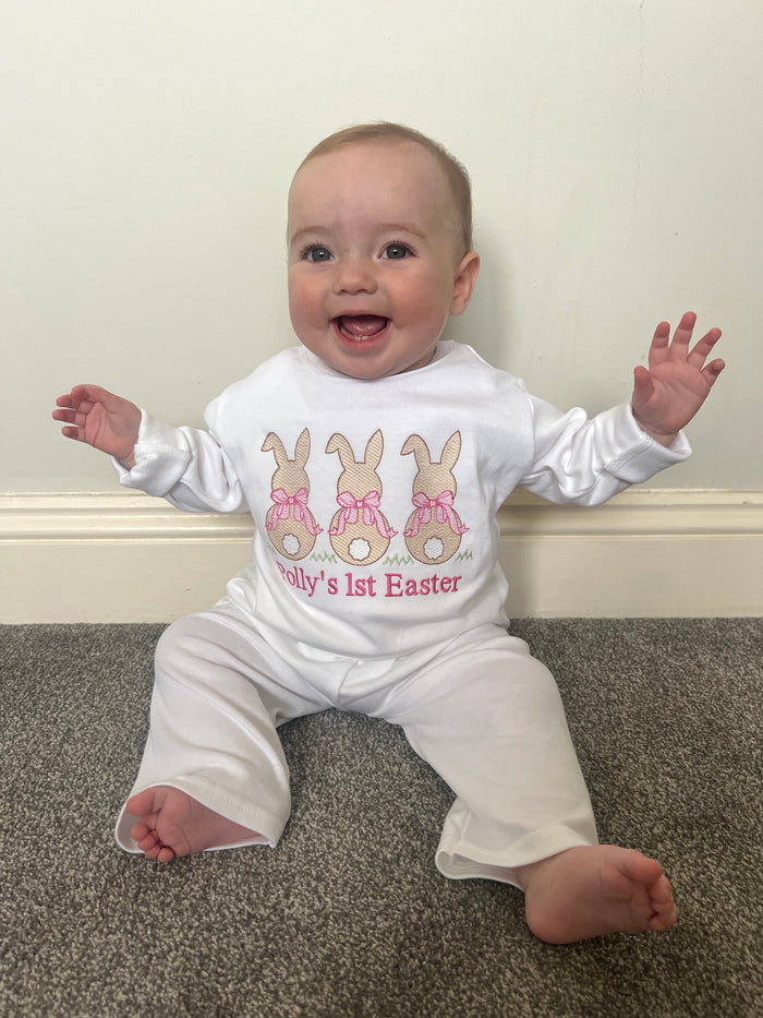 Baby wearing a 'My 1st Easter' onesie on a carpeted floor. 3 bunny’s with pink bows