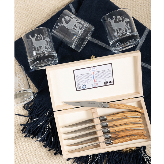 Set of four clear rocks glasses with etched deer design beside wooden-handled steak knives in box on dark fabric.