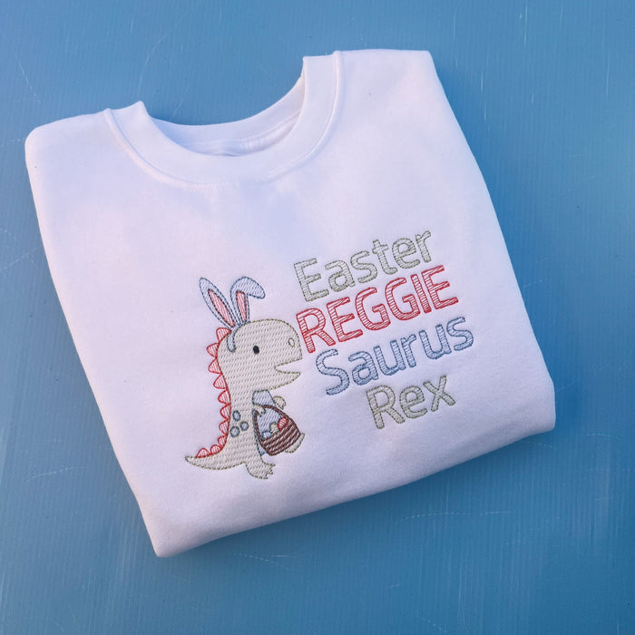 White sweatshirt with a dinosaur with bunny ears and a Easter egg basket with the words Easter Reggie Saurus Rex