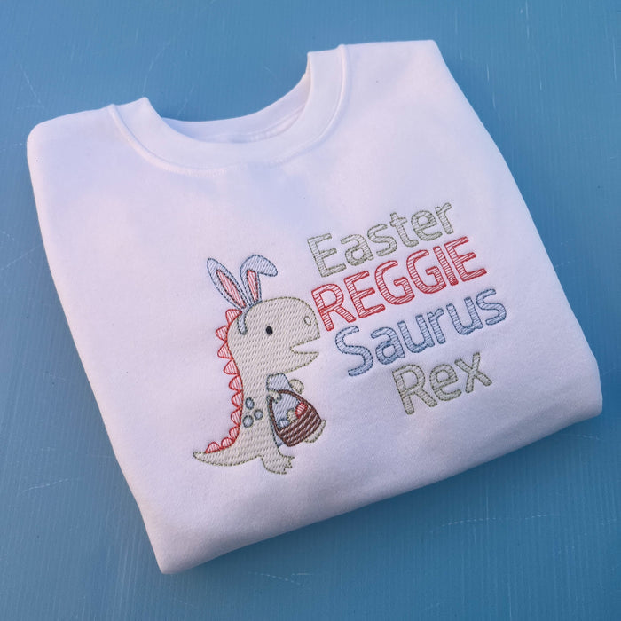 White sweatshirt with a dinosaur with bunny ears and a Easter egg basket with the words Easter Reggie Saurus Rex