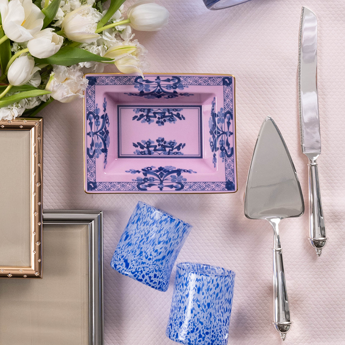 Silver cake knife and server with ornate handles on pink textured cloth beside blue and white marbled tumblers and square.