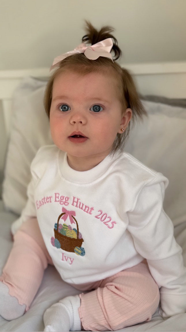 Baby wearing a white shirt with 'Easter Egg Hunt 2023' text and graphics, sitting on a couch.