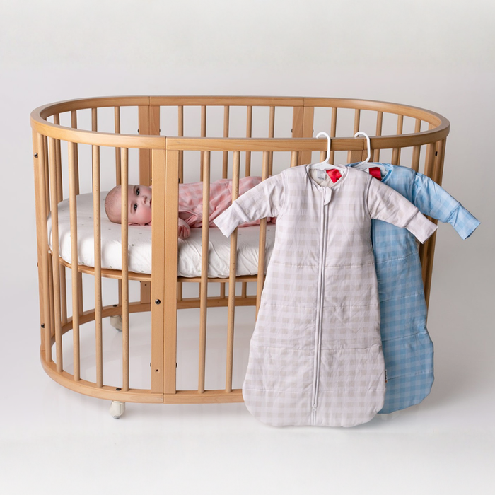 Baby in a wooden cot beside linen grey and blue gingham infant sleep sacks hanging on the rail