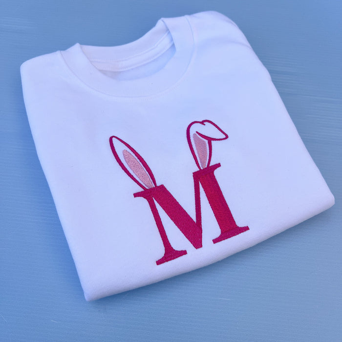 Folded white t-shirt with pink 'M' design with bunny ears on a blue background
