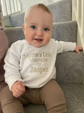 Baby wearing a white sweatshirt with 'Mumma's Little Valentine Jasper' text, sitting on a staircase.