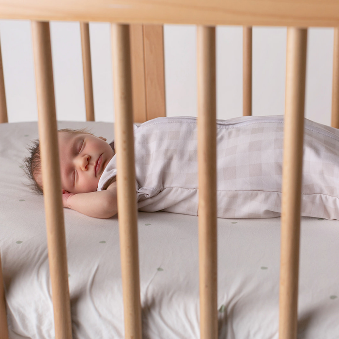 Newborn sleeping soundly in a cot wearing the linen grey gingham newborn swaddle sack