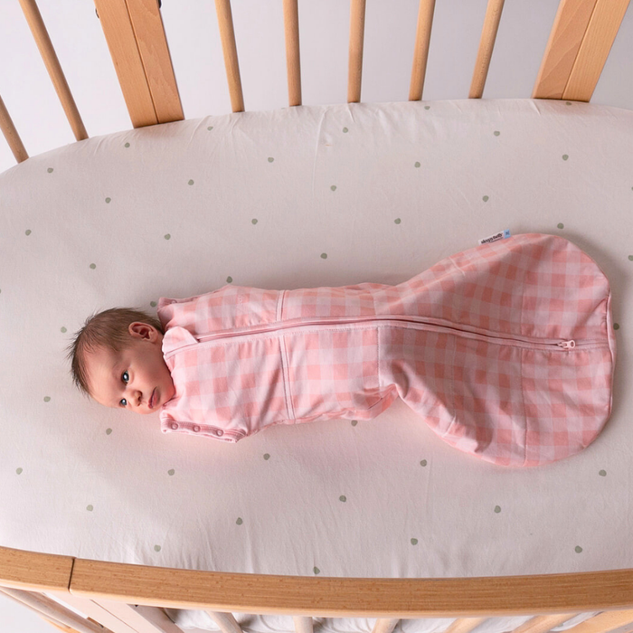 Baby lying in a cot swaddled in the pink gingham newborn swaddle sack with arms tucked in
