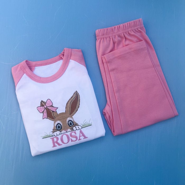 Pink pyjamas with a bun peaking over some grass with a bow on her ear. Finished off with the name Rosa.