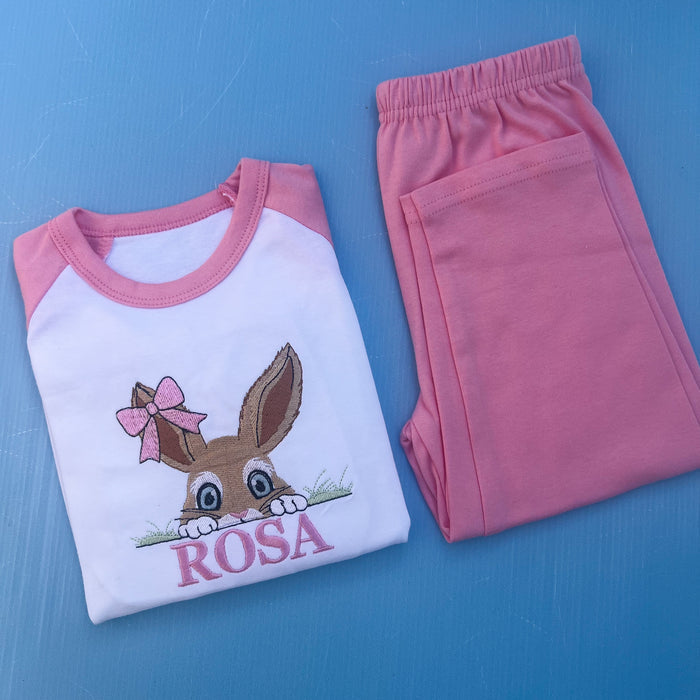 Pink pyjamas with a bun peaking over some grass with a bow on her ear. Finished off with the name Rosa.