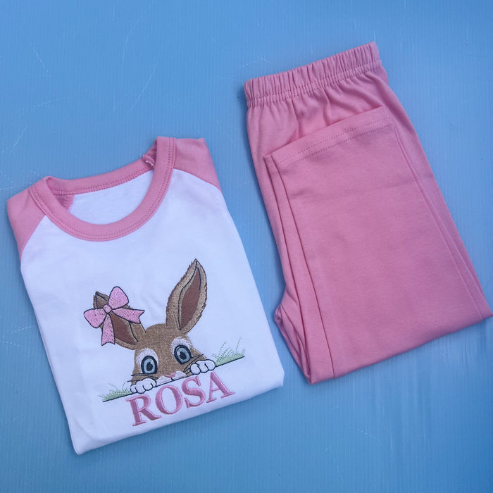 Pink pyjamas with a bun peaking over some grass with a bow on her ear. Finished off with the name Rosa.