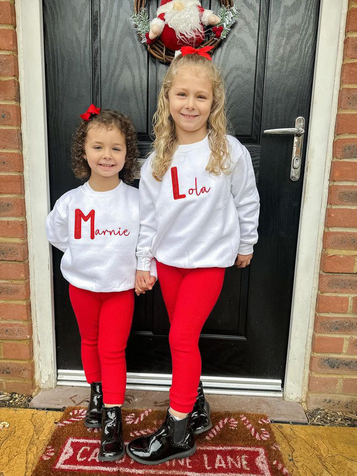 2 girls wearing the red glitter initial sweatshirt stood in front of a door