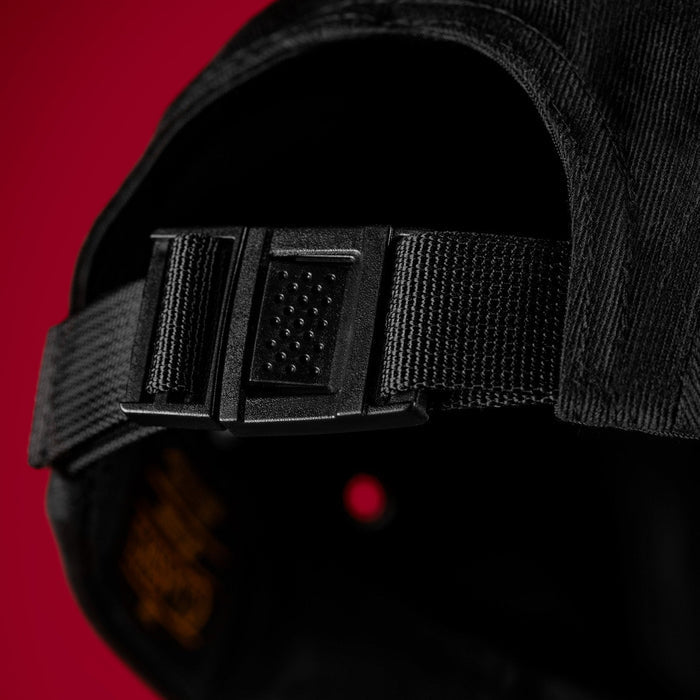 Close-up of a black baseball cap showing the adjustable rear strap and black plastic buckle, set against a solid red background.