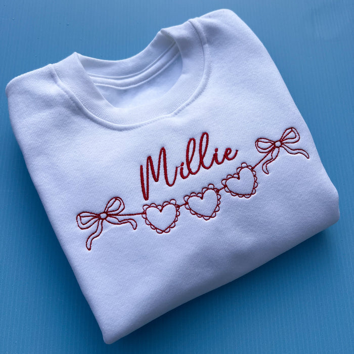 White sweatshirt with the name 'Mille' embroidered in red and a string of hearts below it.