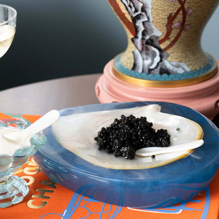 White shell mini spoon resting on blue agate plate with black caviar and cream on side.