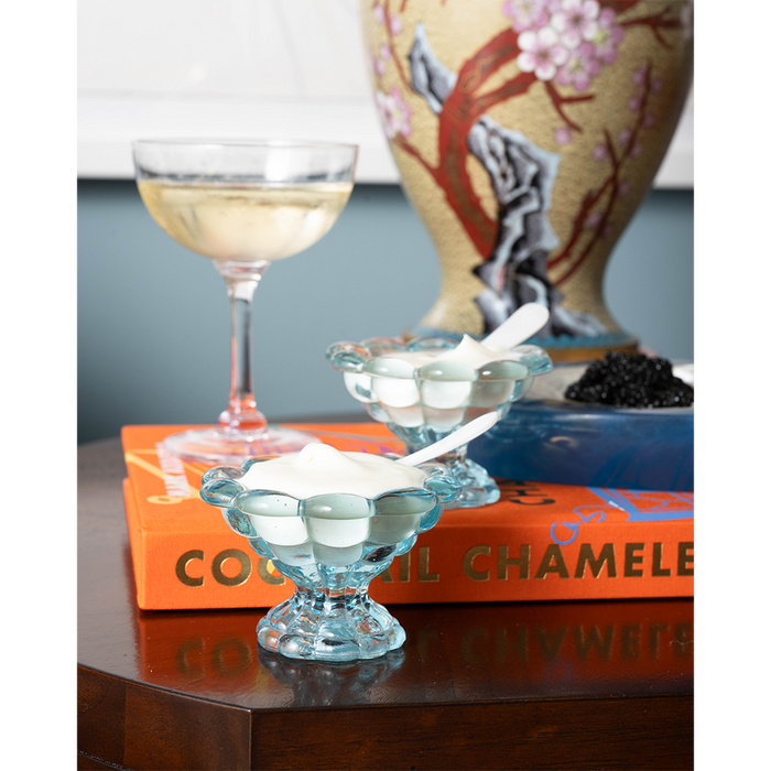 White shell-shaped glass spreader resting on clear footed glass dish on wooden table with books and cocktail glass in.